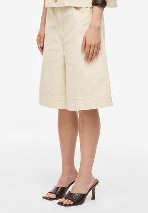 Žena nosí béžové široké šortky, ktoré sú kombinované s tmavohnedými vysokými open-toe mules a širokým hnedým náramkom na zápästí.