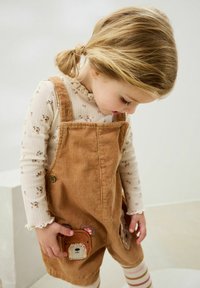 Pettorine in corduroy marrone con due tasche decorate con motivi di orsi. Maglietta a maniche lunghe color crema con motivo floreale e scollatura arricciata. Leggings a righe.