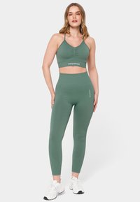 Zelen modrček za šport z V-izrezom in nabranim sprednjim delom, v kompletu z visokimi zelenimi legicami. Obe izdelki sta narejeni iz gladkega, raztegljivega materiala.