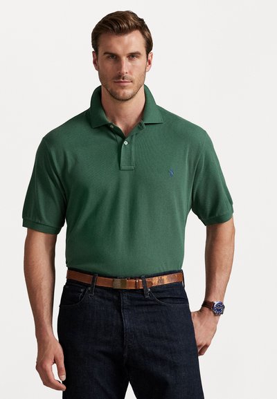 Grüne Polo-Shirt, kurze Ärmel, mit Kragen. Besitzt ein blaues Emblem und wird mit einer dunklen Jeans und einem braunen Ledergürtel kombiniert.