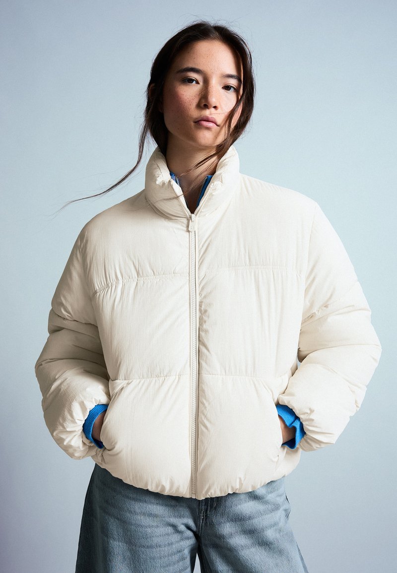 Jeune femme aux cheveux foncés portant une veste matelassée crème sur une chemise bleue, les mains dans les poches, debout devant un fond clair uni.
