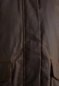 Veste en toile marron foncé avec une surface texturée, dotée d'une couture centrale et de deux poches avant. Détails de couture remarquables le long des bords.