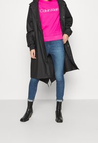 Zwarte lange regenjas over een felroze sweatshirt met het "Calvin Klein"-logo, gecombineerd met een blauwe spijkerbroek en zwarte enkellaarsjes. Textuur: glad, casual pasvorm.