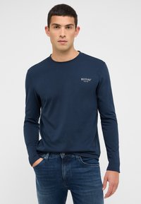 Tumši zils garām piedurknēm krekliņš no kokvilnas. Apaļš kakla izgriezums, pieguļošs siluets ar mazu baltu "MUSTANG" logotipu kreisajā krūtīs. Kombinēts ar zilām džinsa biksēm.