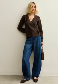 Maglione marrone avvolgente con maniche lunghe, abbinato a jeans in denim blu a gamba larga e scarpe basse marroni. Gli accessori includono una borsa marrone.