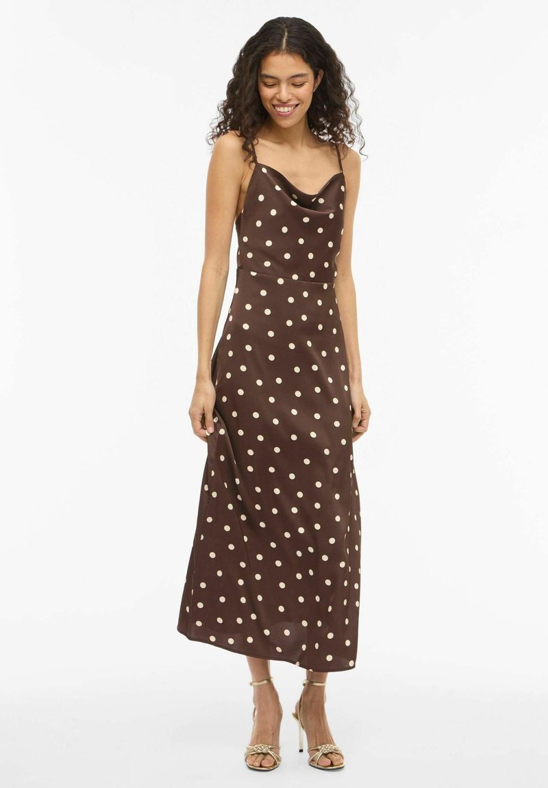 Rjava satenasta obleka z ovratnikom v obliki kapuce, tankimi naramnicami in belimi pikami, ki ima tekočo silhueto in dolžino do sredine meč. Dopomorejo ji sandali z naramnico.