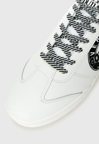 Biele tenisky s čierno-bielymi pruhovanými šnúrkami a okrúhlym čiernym logom na boku s nápisom "CONVERSE."