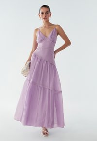 Langes, fliederfarbenes Chiffonkleid mit dünnen Trägern, V-Ausschnitt, gestuftem Design, Perlenakzenten, kombiniert mit einer gewebten Clutch und weißen Riemchensandalen.