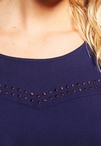 Haut bleu marine avec un col rond et une rangée horizontale de clous en métal sur la poitrine. Texture de tissu lisse.