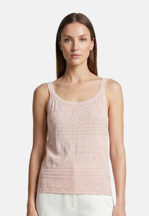 Femme aux longs cheveux lisses portant un haut tricoté sans manches de couleur rose clair et un pantalon blanc, face à un fond uni.