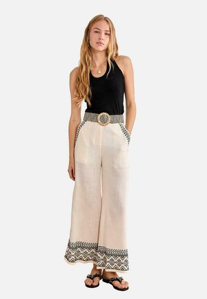 Joven mujer con top negro sin mangas, pantalones anchos beige con bordado negro, cinturón tejido y sandalias negras, de pie frente a un fondo liso.