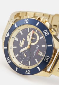 Reloj Lacoste con tono dorado que presenta un bisel con acento azul marino, pantalla de hora dual con tres esferas y una correa de metal texturizada.