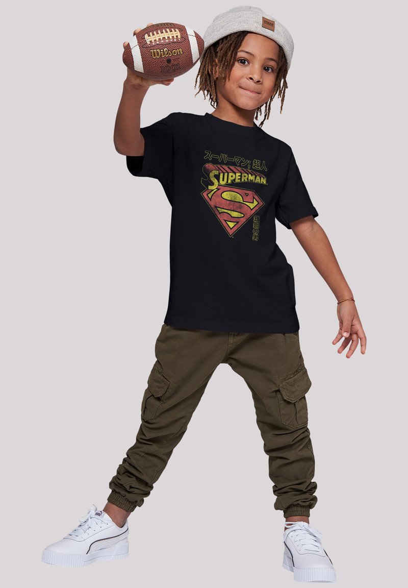 Camiseta negra con un logo de Superman y texto en japonés, combinada con pantalones cargo oliva y zapatillas blancas, sosteniendo un balón de fútbol.
