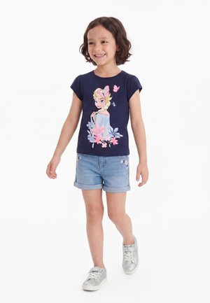 Mädchen lächelt und läuft, trägt ein dunkelblaues T-Shirt mit Frozen-Motiv, helle Jeansshorts mit Blumenstickerei und silberne Sneakers.