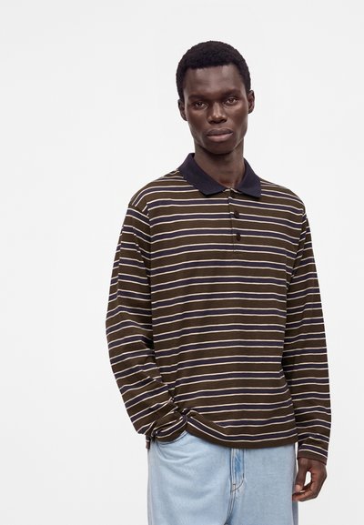 Jeune homme portant un polo rayé marron et blanc à manches longues et un jean bleu clair, debout sur un fond blanc uni.