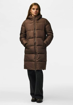 Junge Frau steht und trägt einen langen braunen gesteppte Mantel mit Kapuze, Hände in den Taschen, schwarze Hose und schwarze Schuhe vor einfachem Hintergrund.