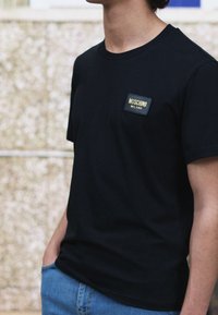 Svart bomullströja med rund halsringning, prydd med en rektangulär Moschino Milano-logotyp i guld och svart på vänster bröst.