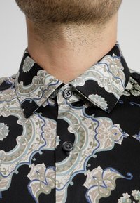 Chemise noire à motifs avec des dessins floraux et géométriques complexes en bleu clair, beige et vert, dotée d'un col classique et d'une fermeture à boutons.