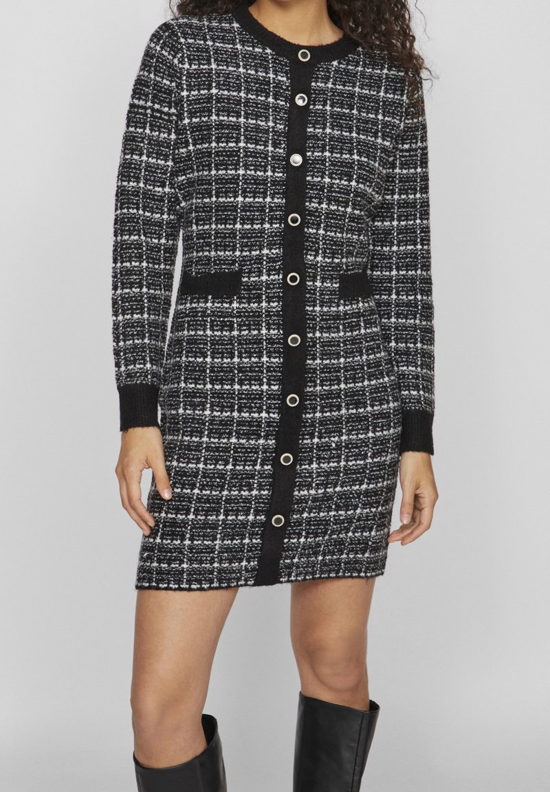 Femme portant une robe tricotée à carreaux noir et blanc avec des manches longues, des boutons sur le devant, deux poches à l'avant et des bottes hautes noires.