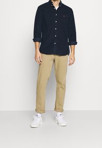 Camicia button-up blu scuro, pantaloni khaki e sneakers bianche. La camicia ha un piccolo logo, maniche arrotolate e un design del colletto classico.