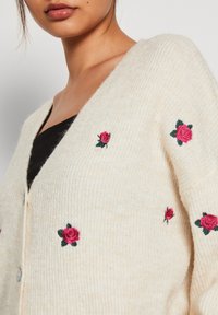 Krämfärgad kofta med texturerad stickning, prydd med rosa rosor broderade över hela framsidan, och med V-ringning.