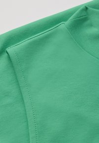 Tessuto di cotone verde con una texture liscia, scollatura arrotondata e dettagli di cucitura visibili lungo i bordi.