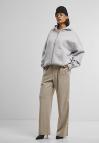 Femme portant un sweat à capuche gris zippé, un pantalon beige à jambes larges, des talons hauts noirs et un bonnet noir, posant devant un fond clair uni.