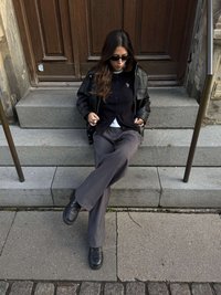 Geacă neagră de piele peste o cămașă neagră cu nasturi, combinată cu pantaloni gri și pantofi negri. Stă pe trepte de piatră, cu o ușă în fundal.