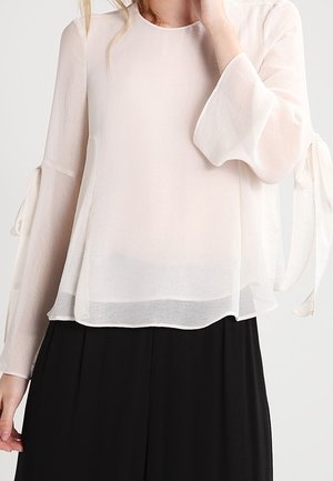 Kvinde iført en gennemsigtig hvid bluse med lange ærmer og bundne manchetter samt løse, sorte bukser med højtaljet pasform, vist fra skuldre til hofter.