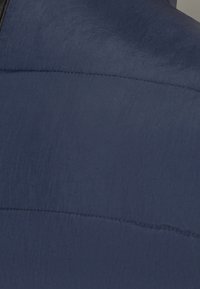 Marineblaues Textil mit sichtbaren Nähten, das eine glatte und leicht glänzende Oberfläche zeigt. Es weist ein geschwungenes Design mit einem schwarzen Akzent auf.