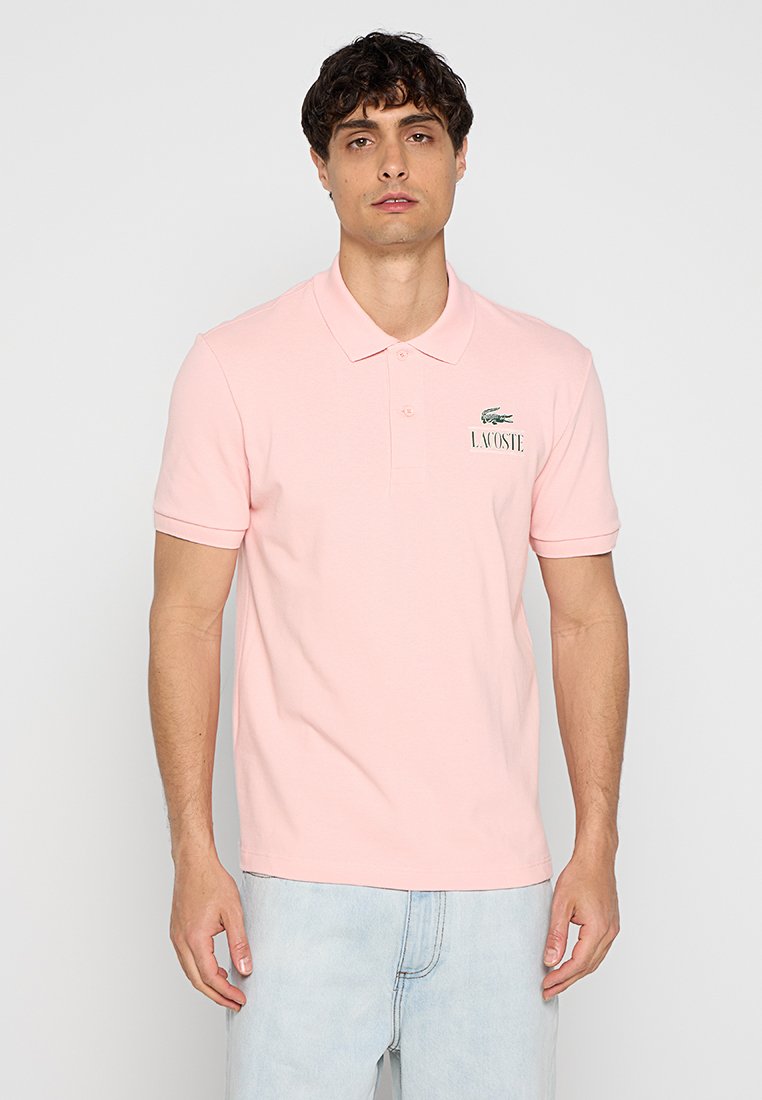 Camisa polo rosa claro hecha de algodón. Presenta un cuello clásico, mangas cortas y un logo de Lacoste bordado en verde en el lado izquierdo del pecho.