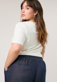 Cremefarbenes Kurzarmoberteil kombiniert mit dunklen Jeanshosen, die einen elastischen Bund und eine Gesäßtasche haben. Weicher Stoff mit einer lockeren Passform.