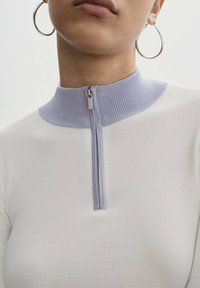 Witte geribbelde zip-up top met een pastelpaarse kraagdetail, voorzien van een metalen rits die tot aan de hals reikt. Strak aansluitend ontwerp.