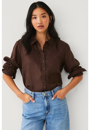 Vrouw met lang zwart haar, gekleed in een donkerbruine blouse met opgerolde mouwen en lichtblauwe, hooggetailleerde spijkerbroek, met handen in de zakken.