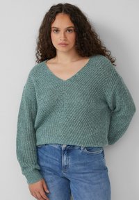 Türkisfarbener, strukturierten Strickpullover mit V-Ausschnitt und langen Ärmeln, kombiniert mit hellblauen Jeans. Besitzt eine lässige, verkürzte Passform und gerippte Akzente.