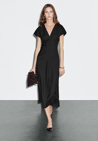 Abito nero midi con scollatura a V e maniche corte, caratterizzato da una silhouette aderente. Abbinato a una pochette scura e tacchi neri.