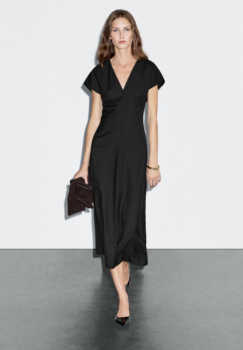 Abito nero midi con scollatura a V e maniche corte, caratterizzato da una silhouette aderente. Abbinato a una pochette scura e tacchi neri.