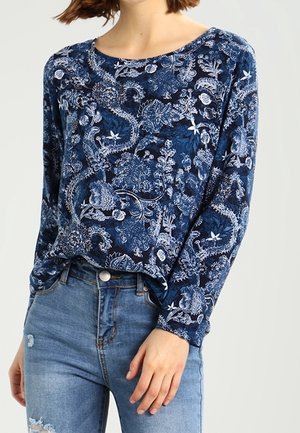 Woman wearing a blue long-sleeve top with white floral and paisley patterns paired with light blue high-waisted jeans.