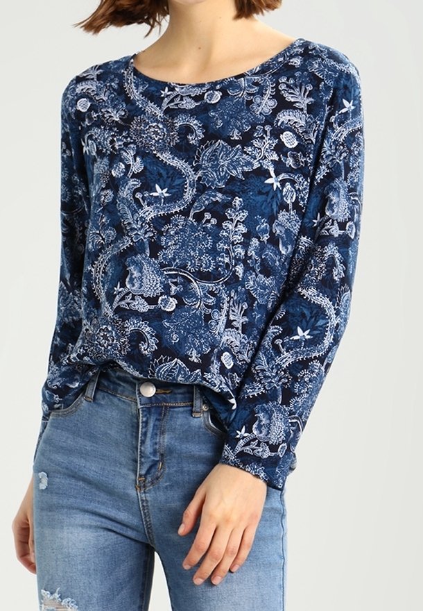 Femme portant un haut bleu à manches longues avec des motifs floraux et cachemire blancs, associé à un jean taille haute bleu clair.