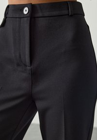 Black tailored trousers made from smooth fabric, featuring a flat waistband, a front seam, and a single button closure.