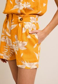 Keltaiset kukkaturasit kevyestä kankaasta, joissa on istuva vyötärö, sivutaskut ja vaalean kermaiset kukkakuviot.