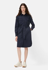 Marineblaues Hemdkleid mit langen Ärmeln, großen Vordertaschen, durchgehender Knopfleiste und einem Taillengürtel. Kombiniert mit hellen Sneakern.