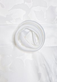 Vit klänning med en texturerad blommönster, försedda med ett satinbälte och en rosett detalj av slätt, vitt tyg.