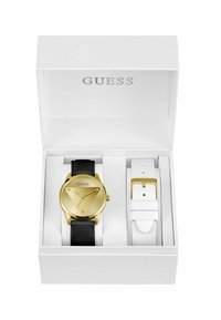 Montre dorée avec bracelet en cuir noir dans une boîte blanche, inclut un bracelet supplémentaire blanc avec boucle dorée. Design minimaliste, cadran épuré.