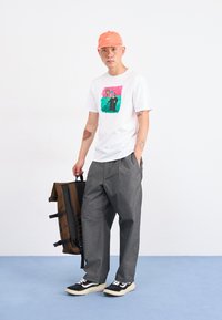 Hvit t-skjorte med fargerik grafikk, grå vide bukser, brun ryggsekk og svarte joggesko med hvite detaljer. Oransje caps på hodet.