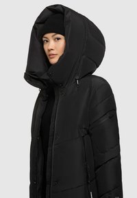 Chaqueta acolchada negra con una gran capucha estructurada, bolsillos con cremallera y una textura suave y brillante. Los acentos incluyen botones a presión y un corte ajustado.