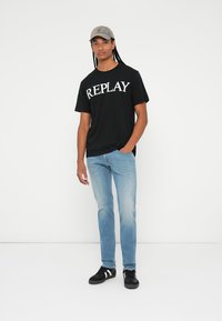 Crna majica s logom "REPLAY" u bijeloj boji, svijetloplave traperice i crne tenisice. Osoba nosi svijetlosivu kapu. Jednostavna, ležerna odjeća.