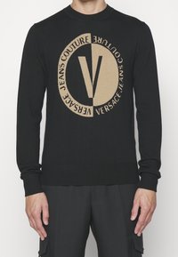 Pull en maille noire avec un grand logo circulaire beige affichant le texte "Versace Jeans Couture" et un "V" prominent au centre.