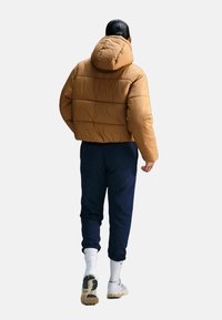Brązowa kurtka puchowa z kapturem, o pikowanej konstrukcji, zestawiona z granatowymi spodniami dresowymi i białymi skarpetkami, widok z tyłu.