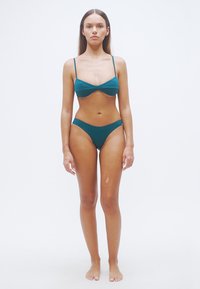 Διπλό μπικίνι σε πετρόλ χρώμα με λεπτές λουρίδες. Λείο ύφασμα με ελάχιστες λεπτομέρειες. Το επάνω μέρος του σουτιέν έχει απλή κοπή και το κάτω μέρος είναι χαμηλοκάβαλο.